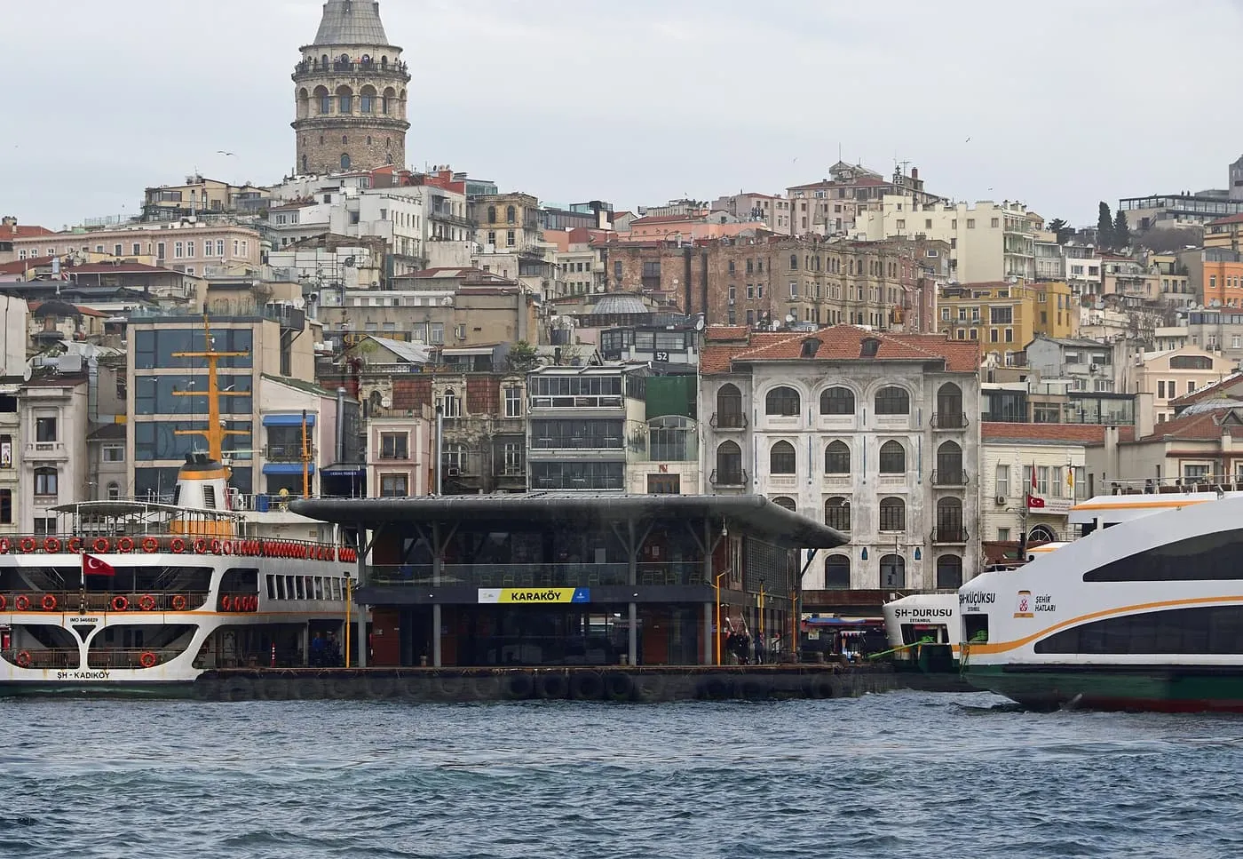 Ferries moored beside the dark-roofed Karaköy ferry pier, with the Galata Tower and tiers of old buildings rising behind