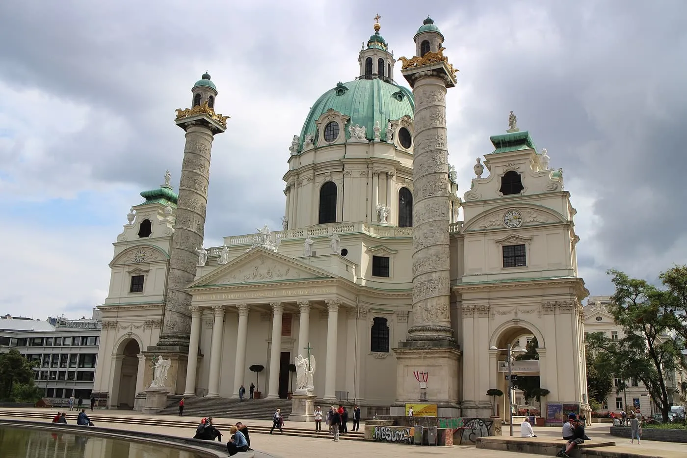 The Karlskirche, a Baroque church with a green copper dome, a columned portico and two tall carved columns, seen across a pool