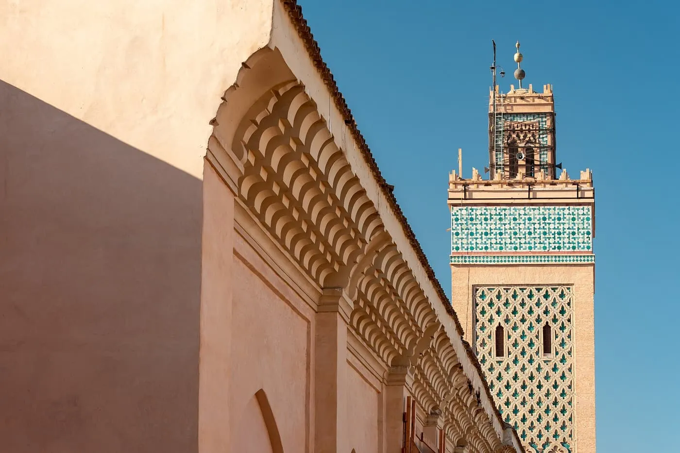 The tiled minaret of the Kasbah Mosque against a blue sky, beside a pink wall with a carved scalloped cornice