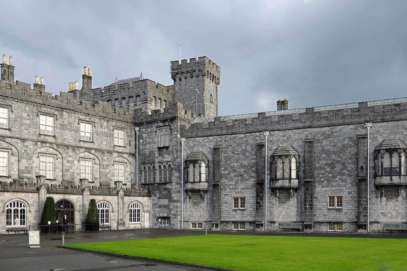 The grey limestone walls and corner tower of Kilkenny Castle seen across a lawn under a stormy sky