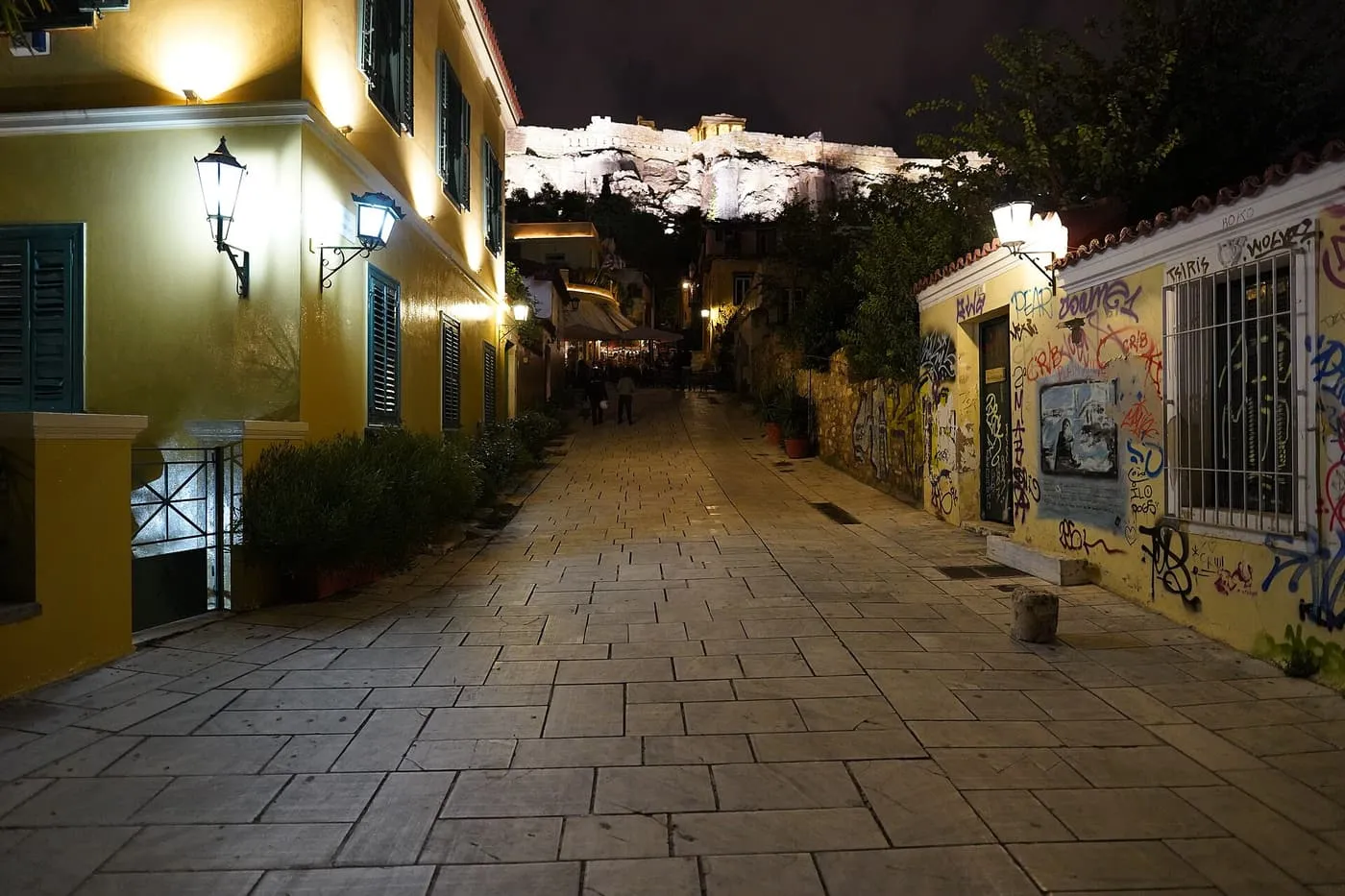 A paved lane in Plaka at night between lit yellow houses, with the floodlit Acropolis wall and the Erechtheion above the rooftops at the end of the street