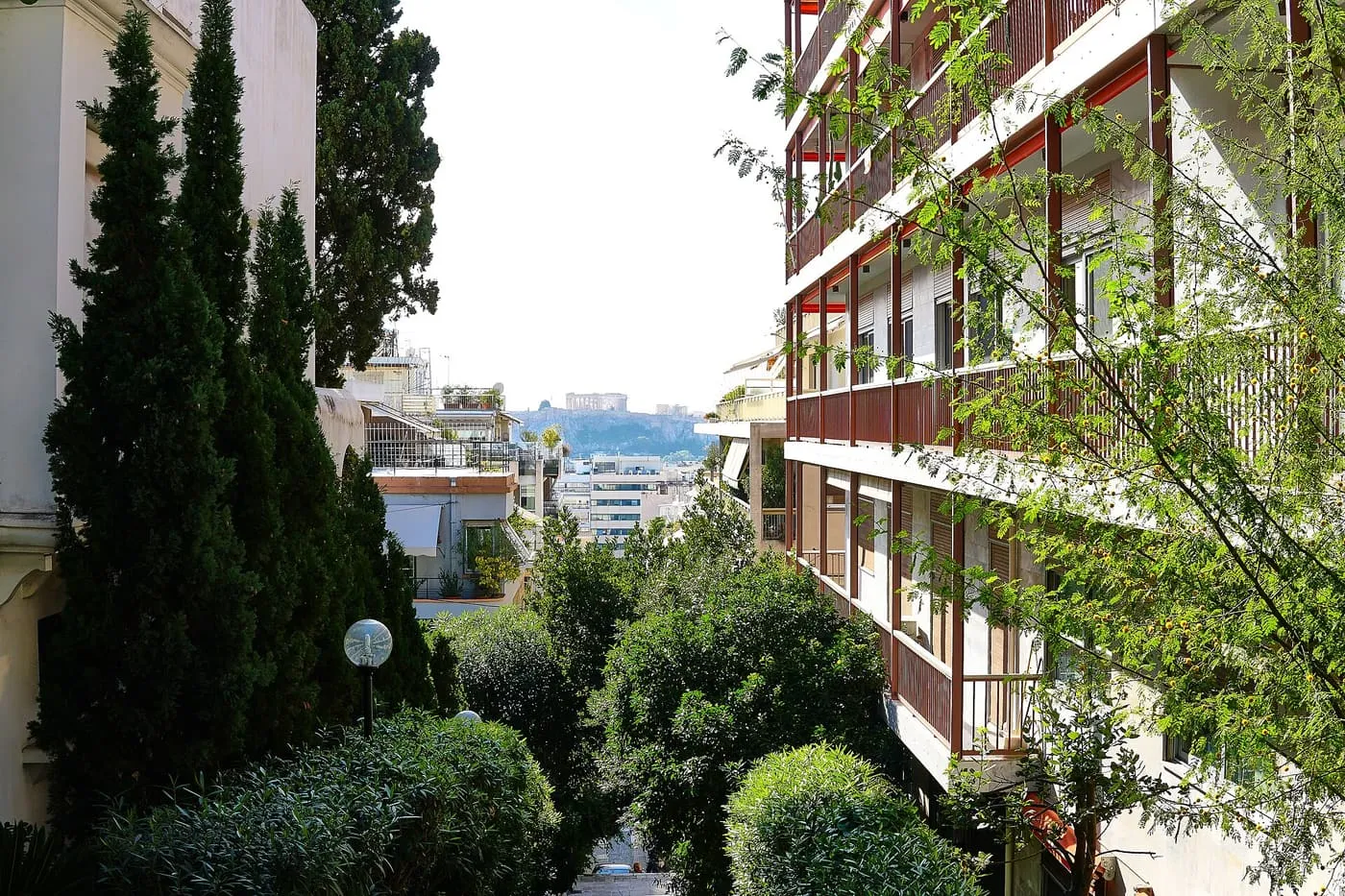 A steep tree-lined street between apartment blocks in Kolonaki, with the Parthenon small on its rock at the far end of the view