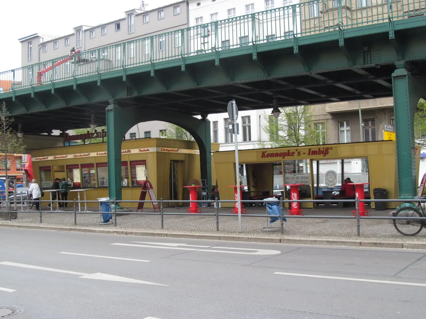 A yellow sausage stand with the sign Konnopke's Imbiss beneath the green steel U2 viaduct on Schönhauser Allee