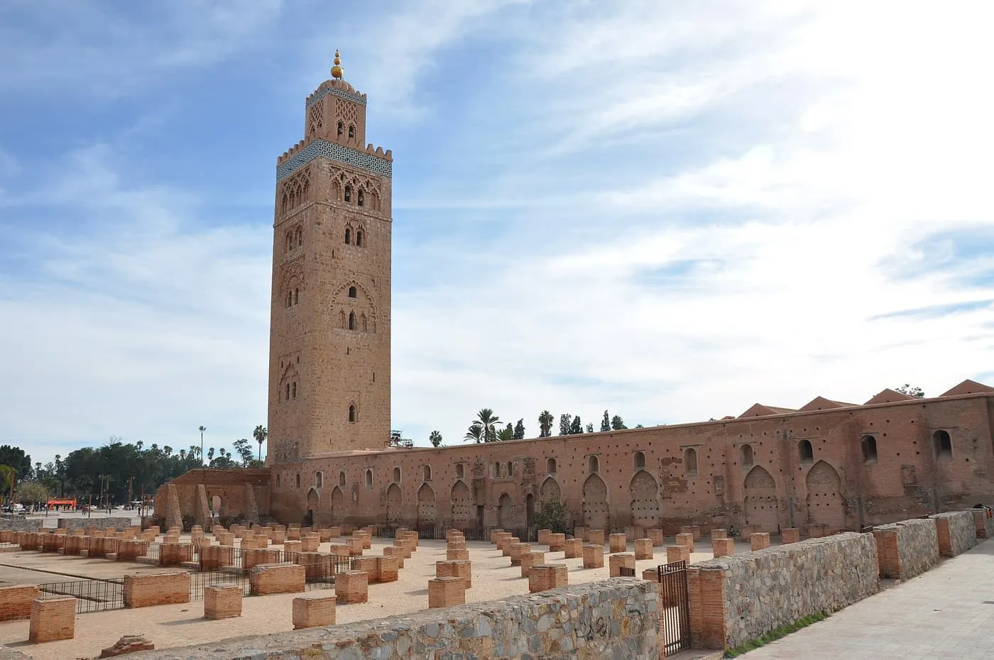 The Koutoubia minaret rising above the long wall of the mosque, with low brick columns marking foundations in front