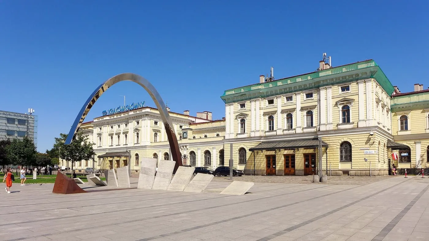 The historic Kraków Główny station building across a paved square with a steel arch sculpture