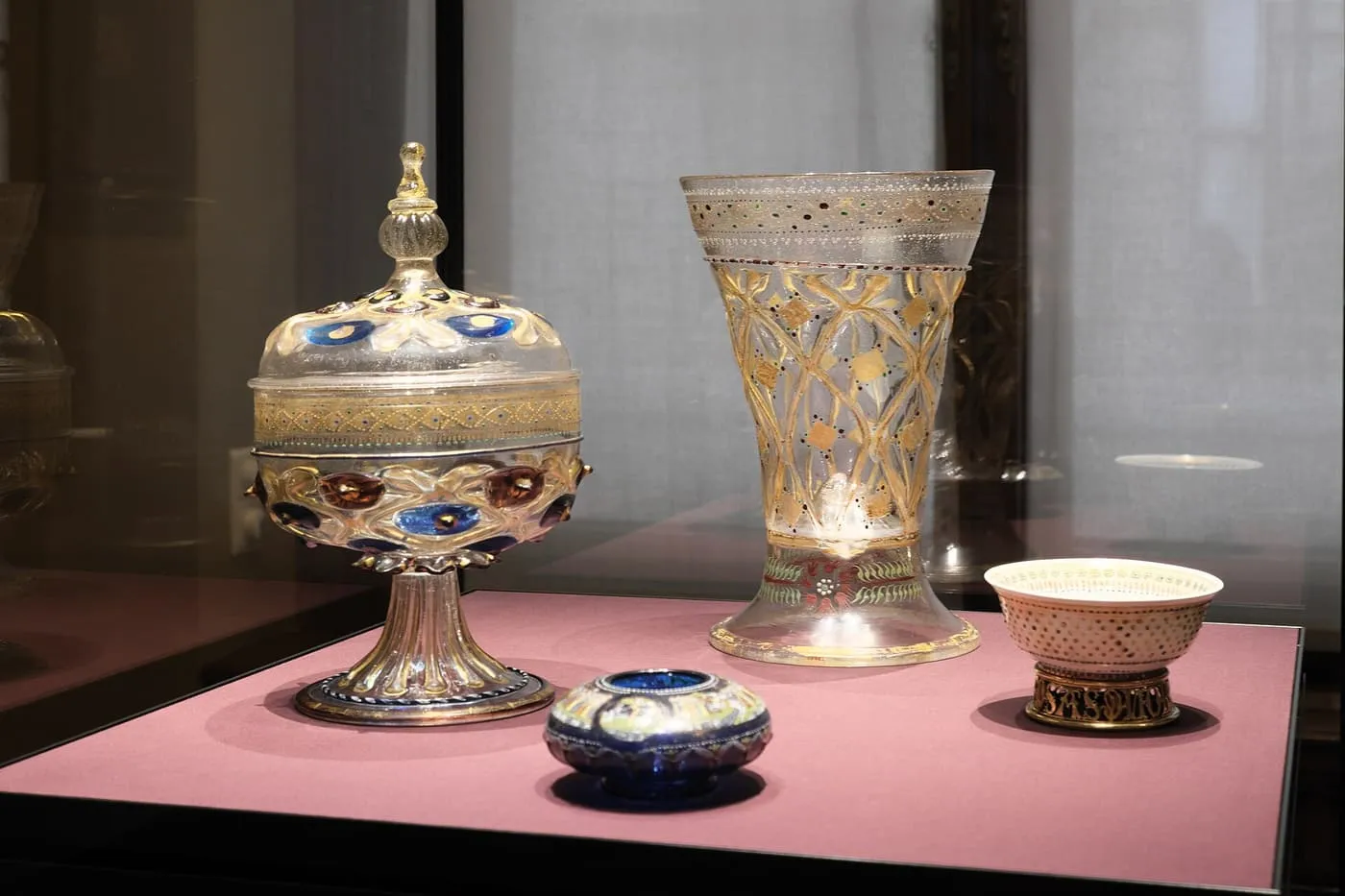 Glass and enamelled vessels in a display case, including a lidded cup on a stem and a tall gilded beaker