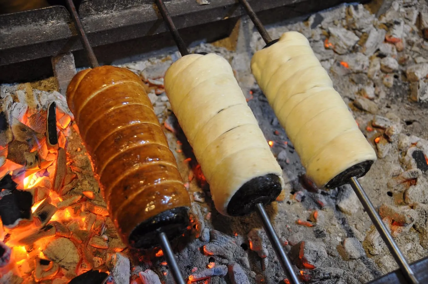 Three chimney cakes on spits turning over glowing embers, one baked golden and two still pale dough