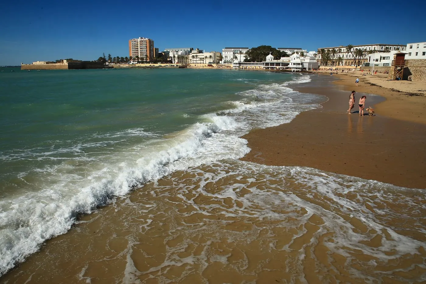 Waves on the sand of La Caleta beach, with the white bathing pavilion and the castle at the far end of the bay