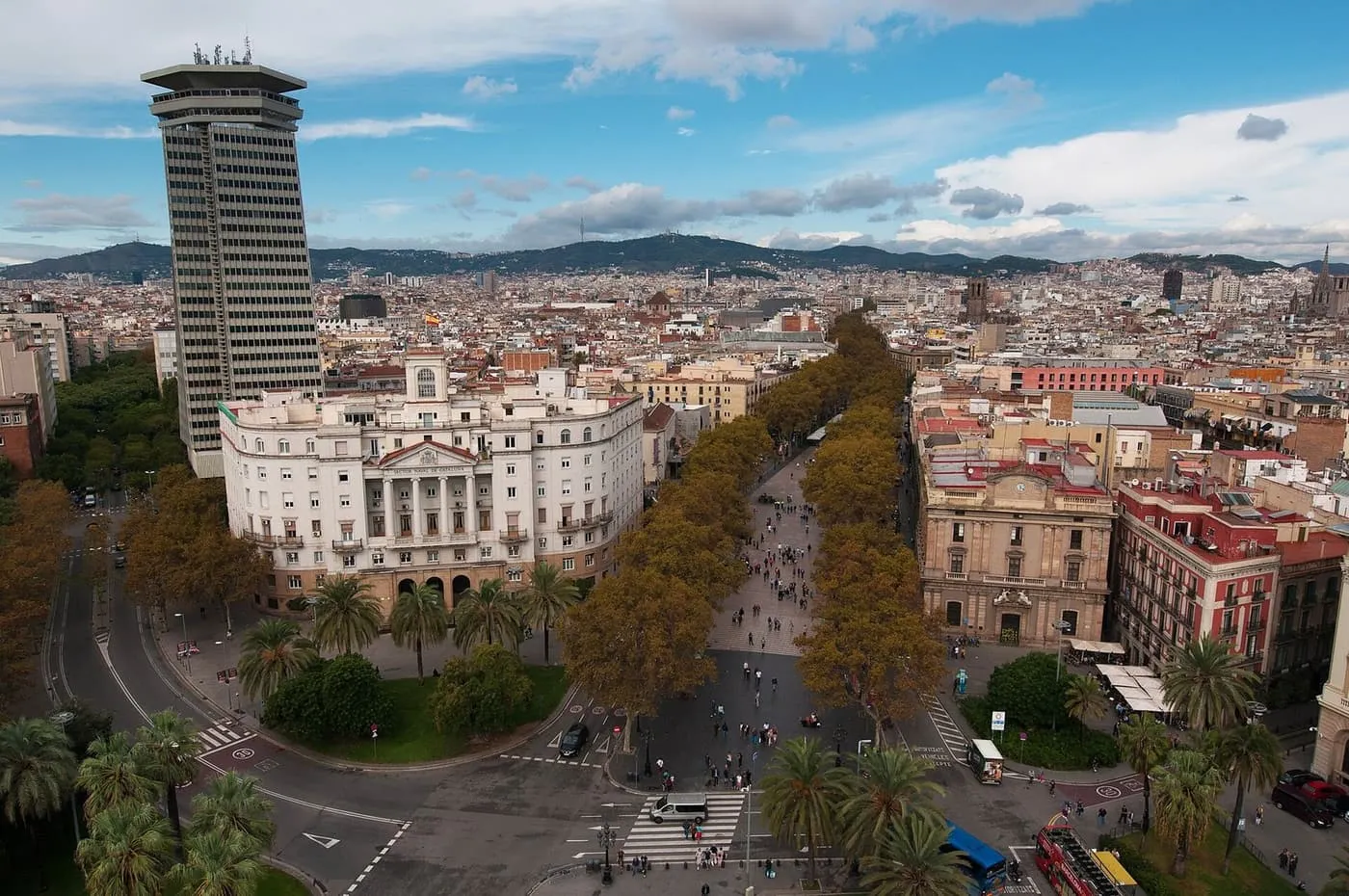 La Rambla seen from above: a tree-lined pedestrian boulevard running between old buildings, with the city and hills behind