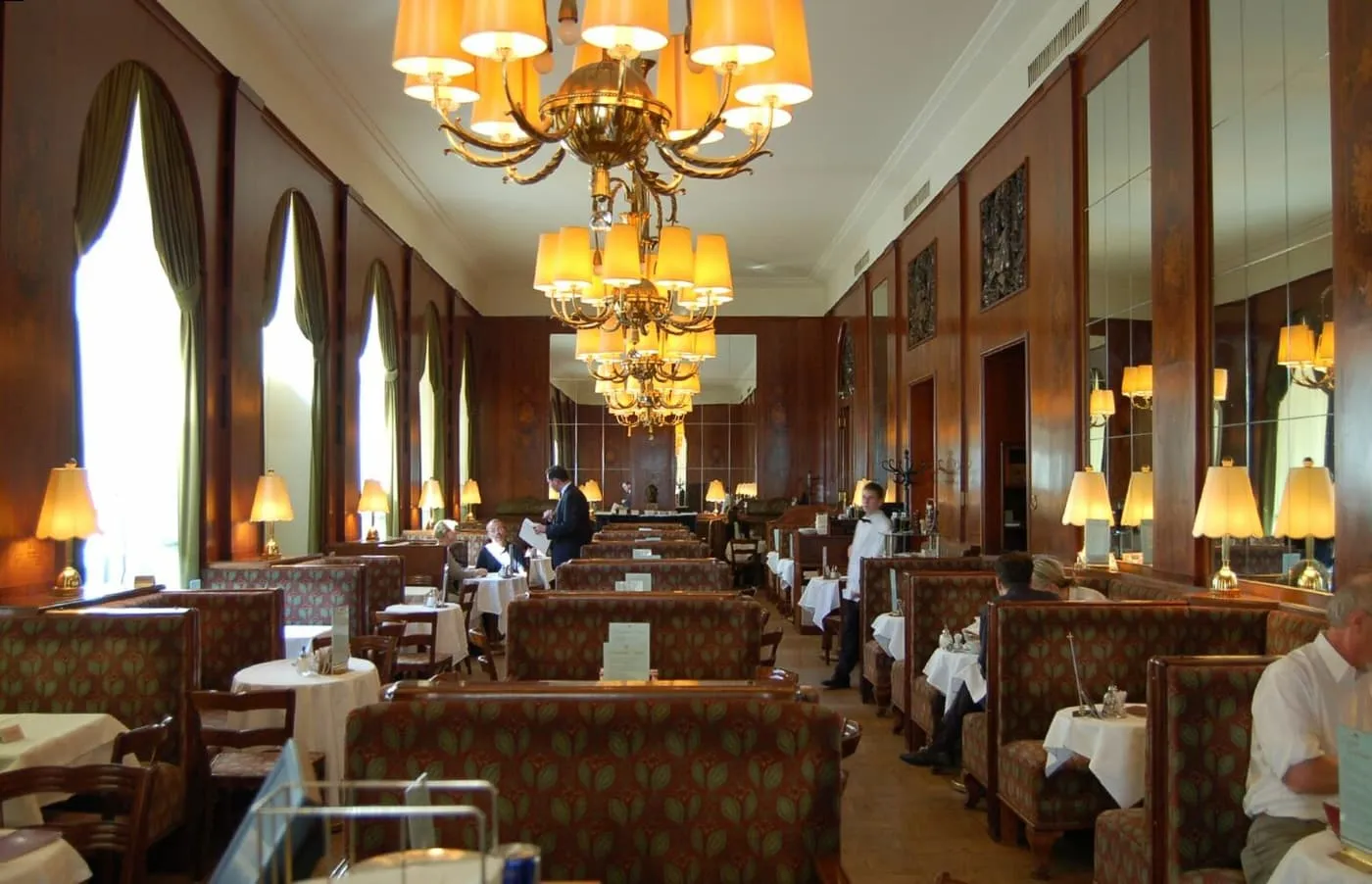 The long panelled room of Café Landtmann, with brass chandeliers, tall windows and rows of upholstered booths and white-clothed tables