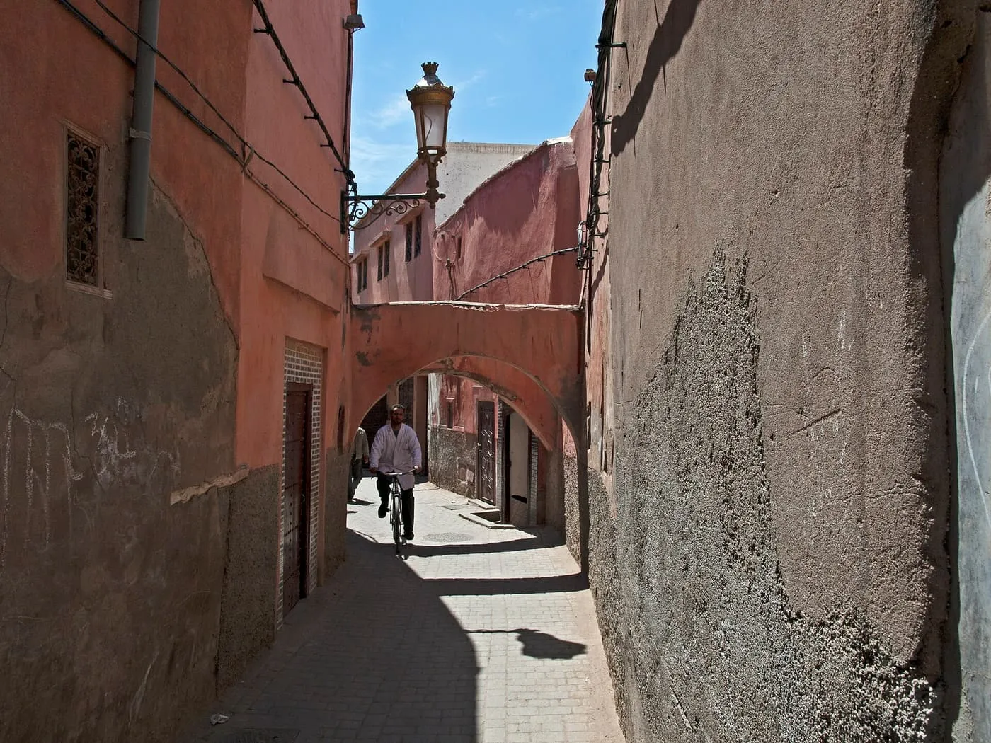 A narrow pink-walled lane with a street lamp and an arch over it, and a cyclist in the distance