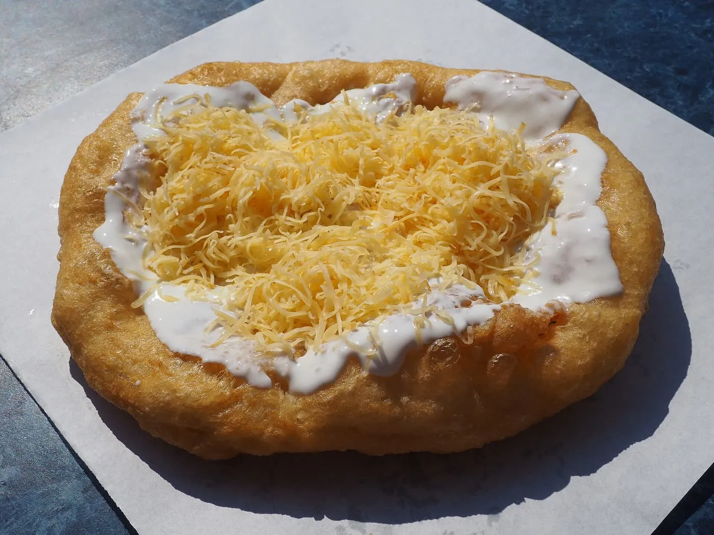 A fried lángos flatbread topped with sour cream and grated cheese on a sheet of paper