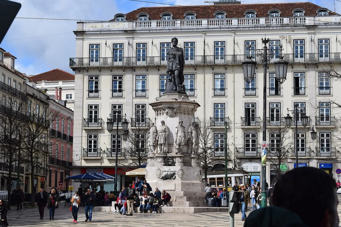 The monument to Luís de Camões in Praça Luís de Camões, with cream-coloured buildings and a yellow tram behind