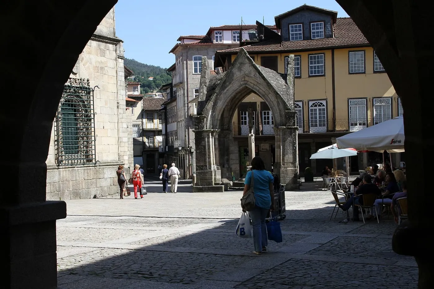 Largo da Oliveira in Guimarães seen through an arch: a cobbled square with a Gothic stone canopy, café umbrellas and old townhouses
