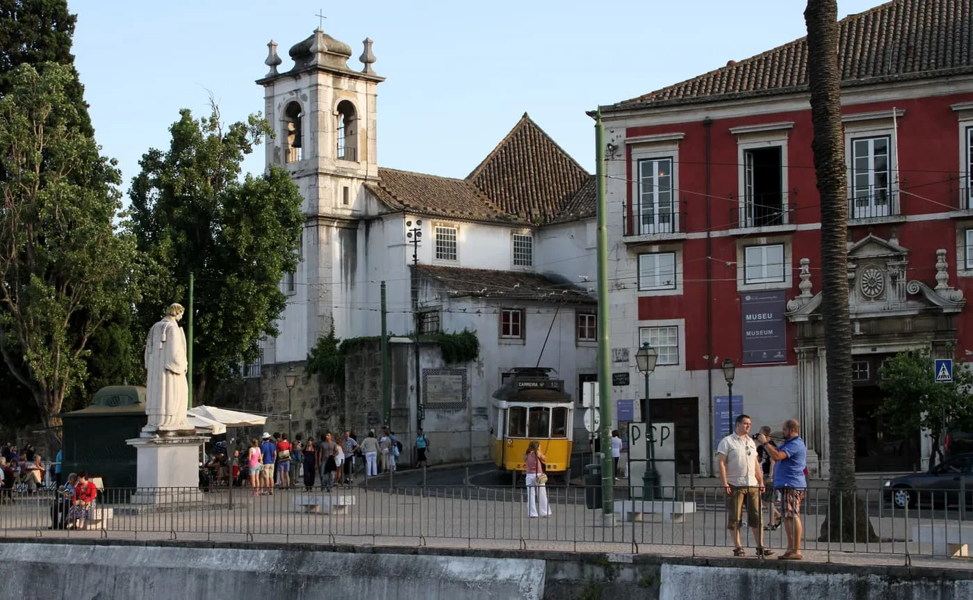 Largo das Portas do Sol, a cobbled square with a statue, a yellow tram passing in front of a white church with a bell tower, and a red palace beside it