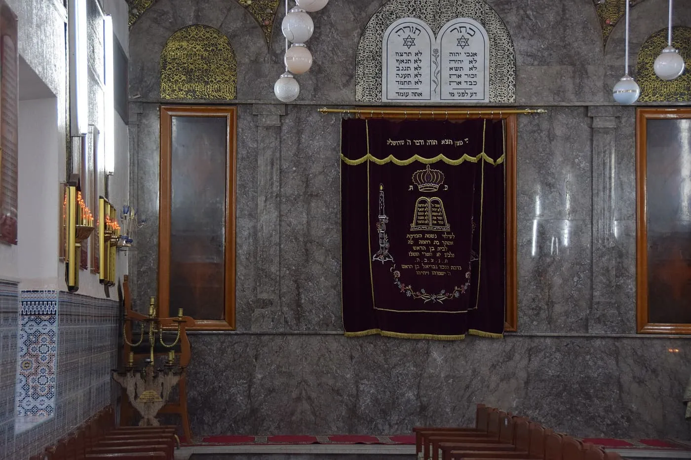 The marble wall of the Lazama Synagogue's prayer hall, with a curtained ark under two stone tablets of the Ten Commandments and tiled walls beside it