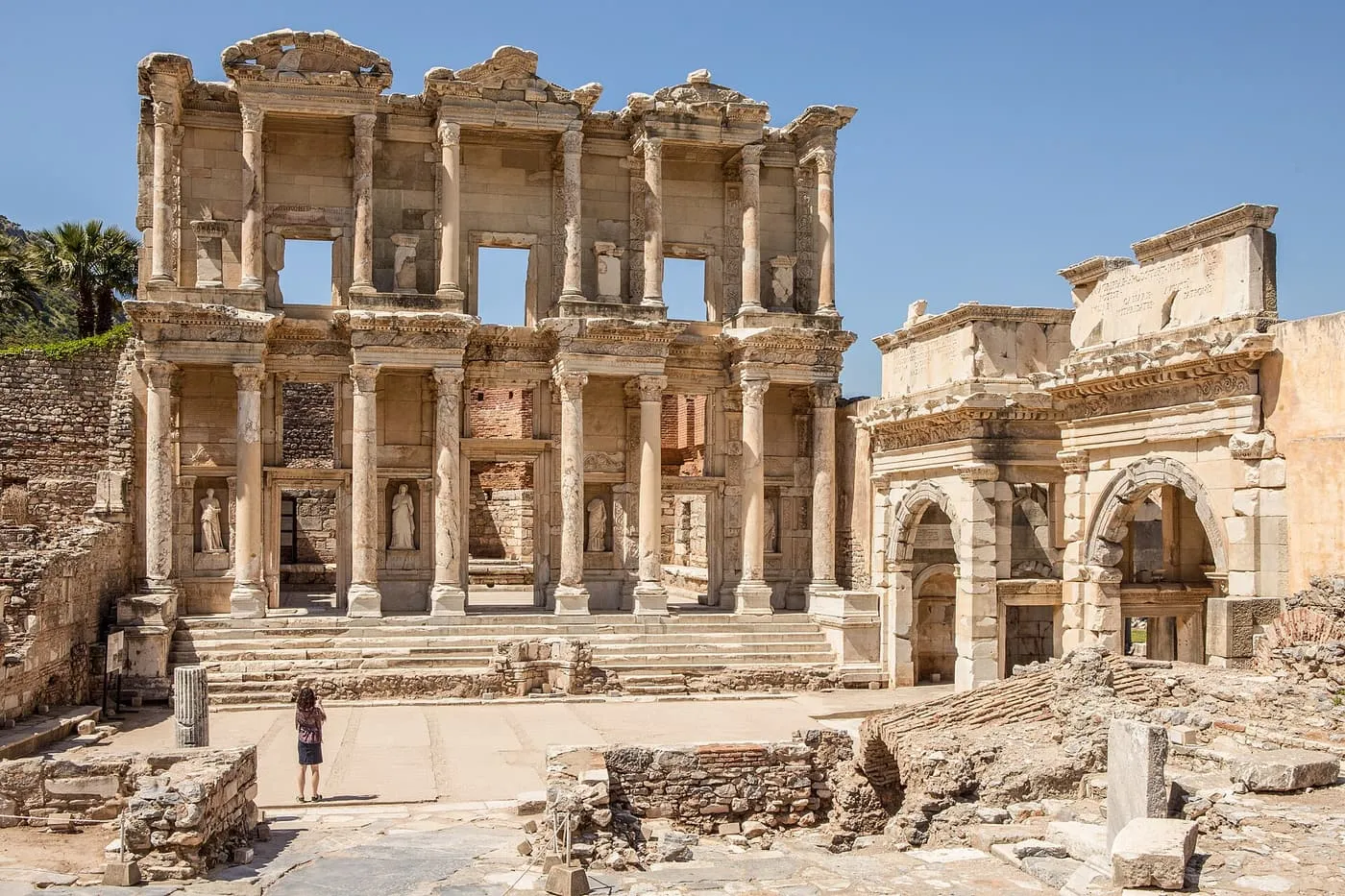 The two-storey columned façade of the Library of Celsus at Ephesus, with a ruined gate beside it and one visitor in front