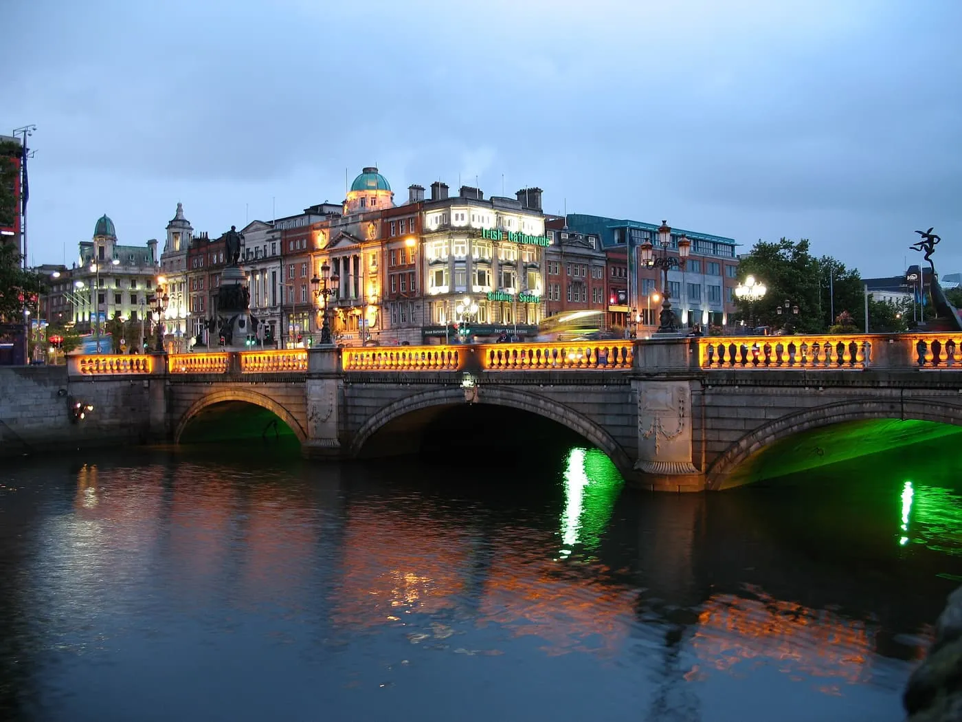 O'Connell Bridge over the Liffey at dusk, its balustrade lit amber and its arches lit green, with buildings along the quay behind