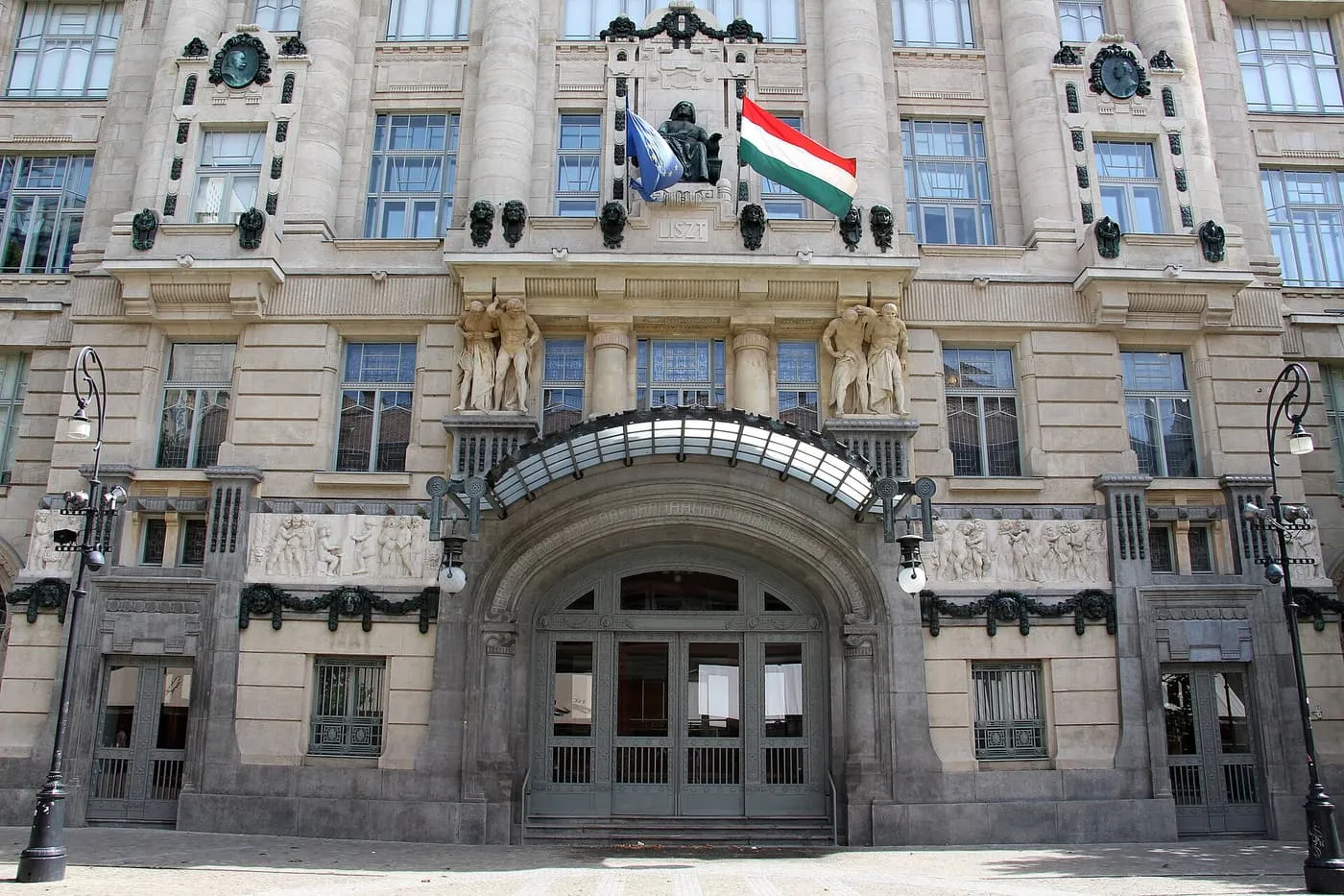 The entrance of the Liszt Academy, a stone Art Nouveau facade with a glass canopy over the doors and a pair of sculpted figures above it