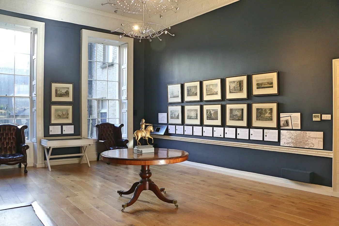 A ground-floor room at the Little Museum of Dublin, with a round table and a small gilt horseman, framed prints on a dark wall and tall sash windows