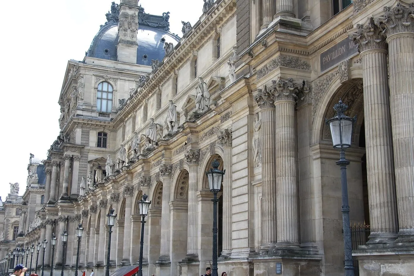 The carved stone facade of the Louvre palace, with statues in niches and a row of lamp posts beside the arcade