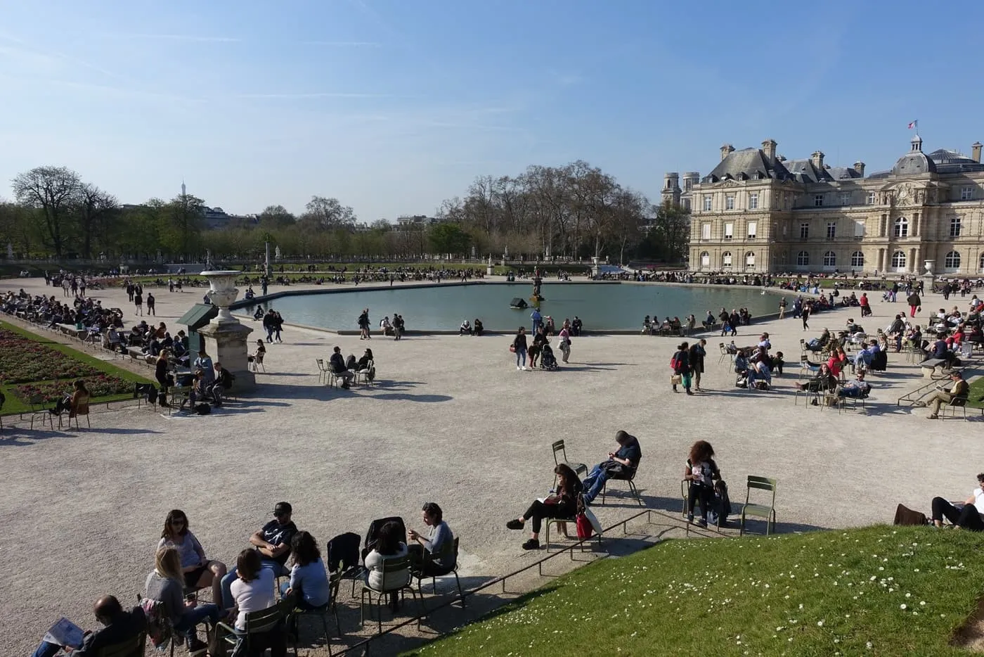 The large basin of the Luxembourg Gardens with the Palais du Luxembourg on the right and people sitting on green metal chairs around the gravel terrace