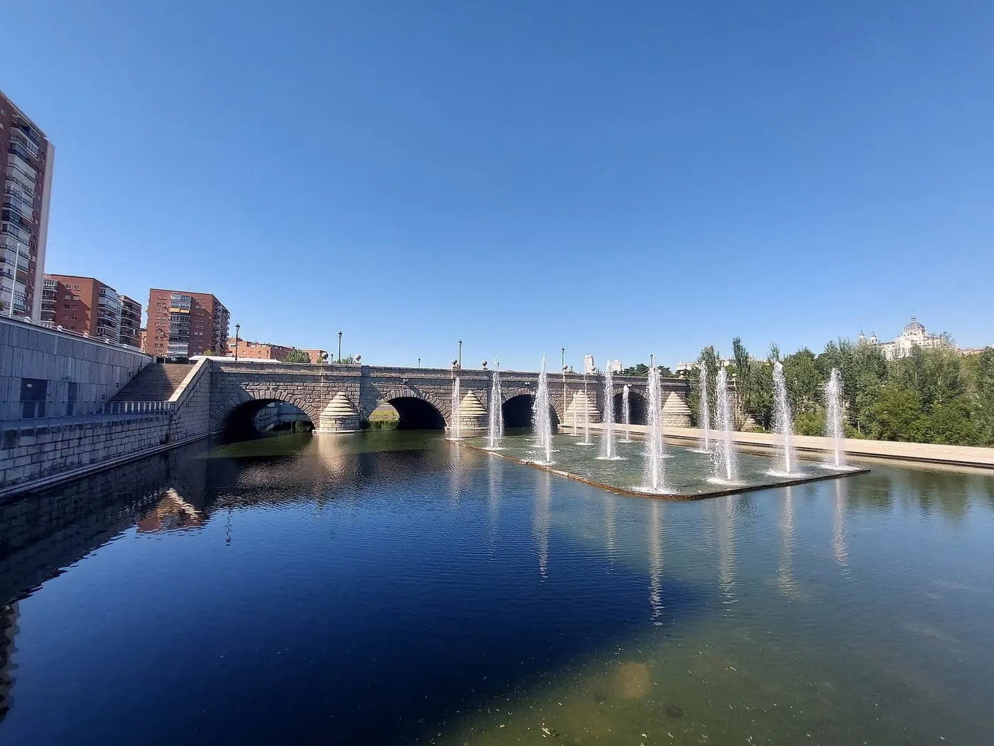 The stone arches of the Puente de Segovia over the Manzanares in Madrid Río, with a row of fountains playing in the water under a clear blue sky