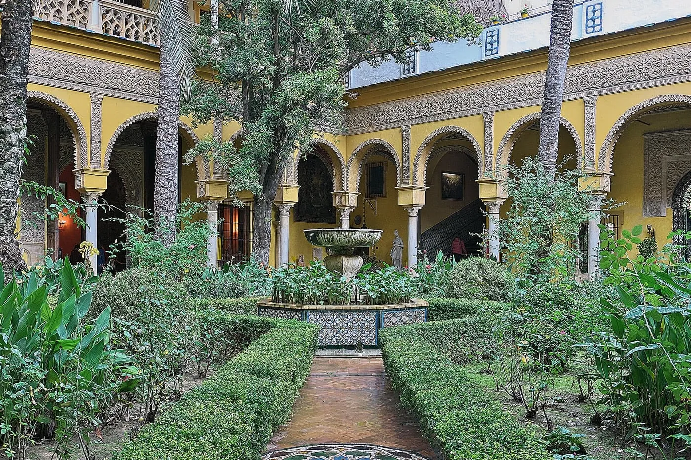 The main courtyard of the Palacio de las Dueñas, with a tiled fountain, clipped hedges and yellow arcaded galleries