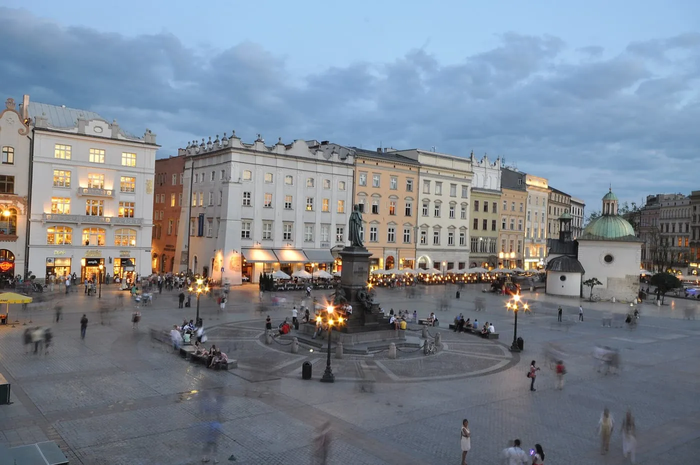 The Main Market Square at dusk, with the Adam Mickiewicz monument, people crossing the square and the small domed St Adalbert's Church on the right