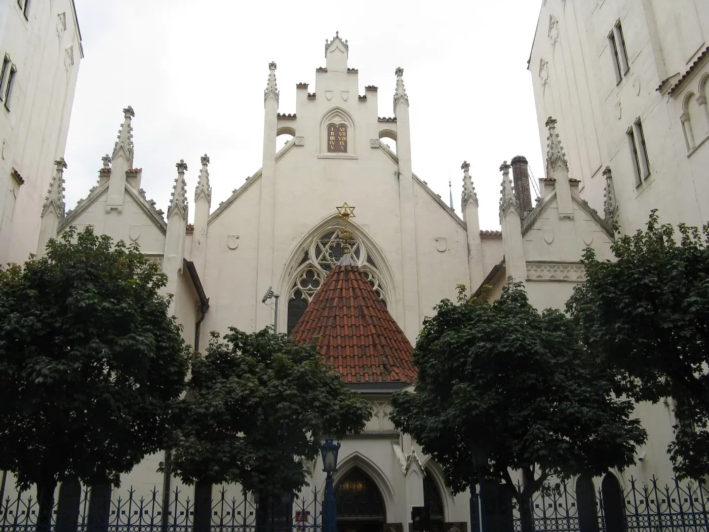 The white neo-Gothic front of the Maisel Synagogue behind trees and an iron fence