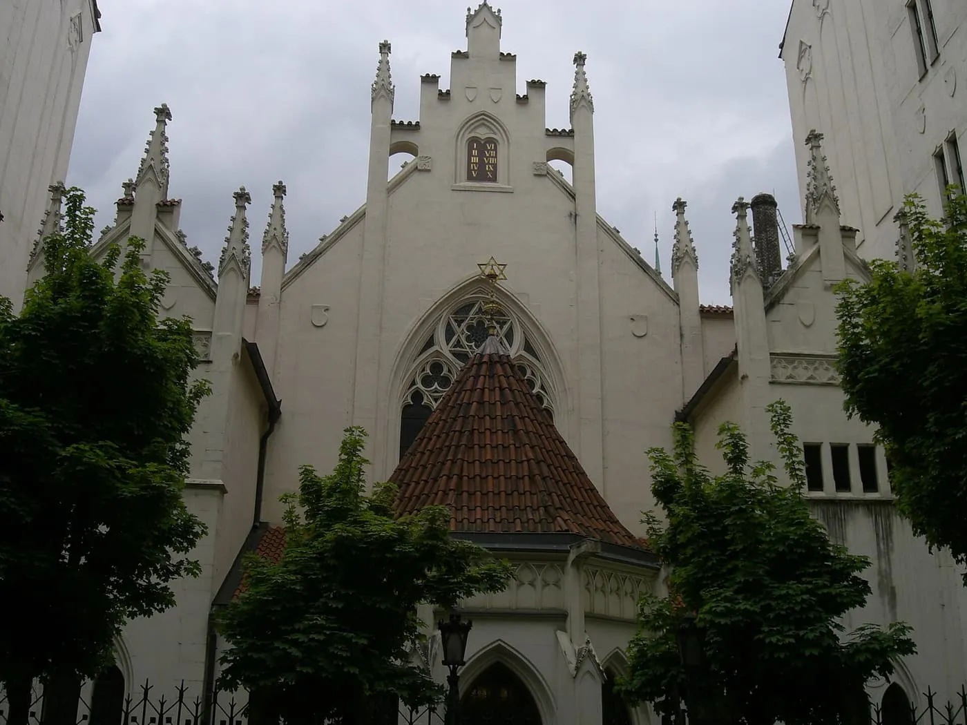 The white neo-Gothic front of the Maisel Synagogue, with pinnacles, a rose window and a red roof, behind trees and an iron fence