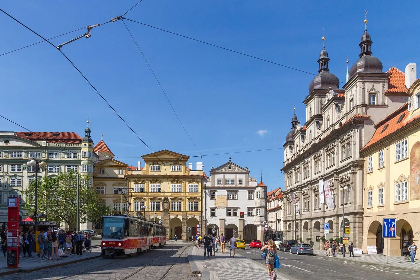 A red and white tram at a stop on Malostranské náměstí, with Baroque palaces and a church with two domed towers around the square