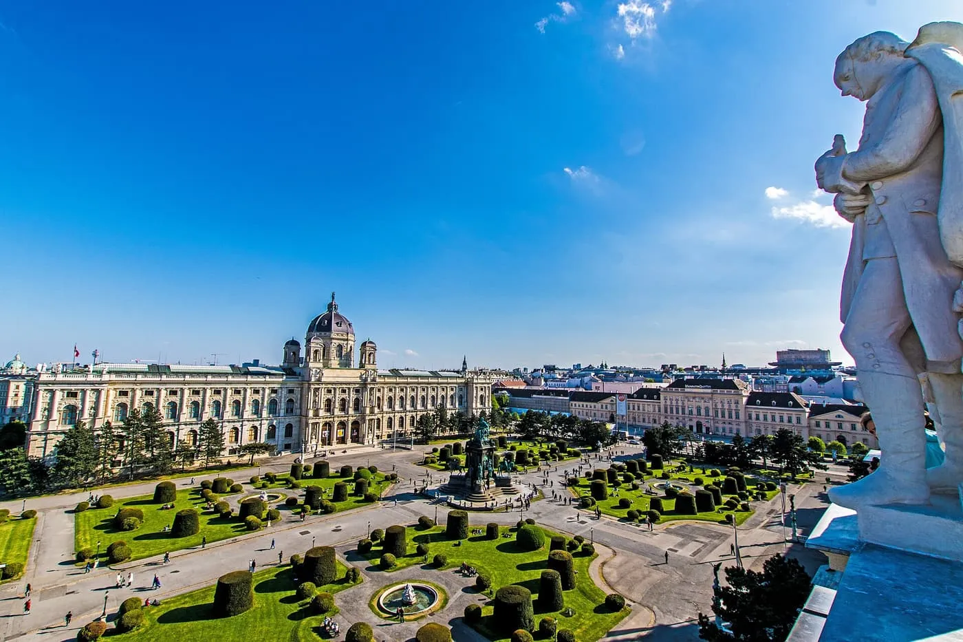 The Kunsthistorisches Museum with its central dome across Maria-Theresien-Platz, seen from above, with clipped hedges below and a statue's shoulder in the foreground