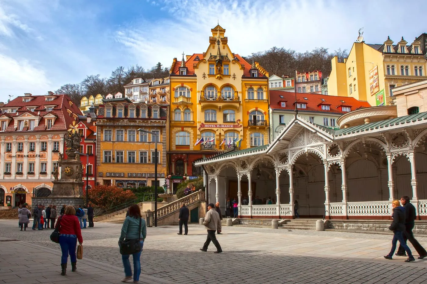The white timber Market Colonnade in Karlovy Vary, with lace-like carved fretwork and a green roof, in front of yellow and pastel townhouses on the hillside