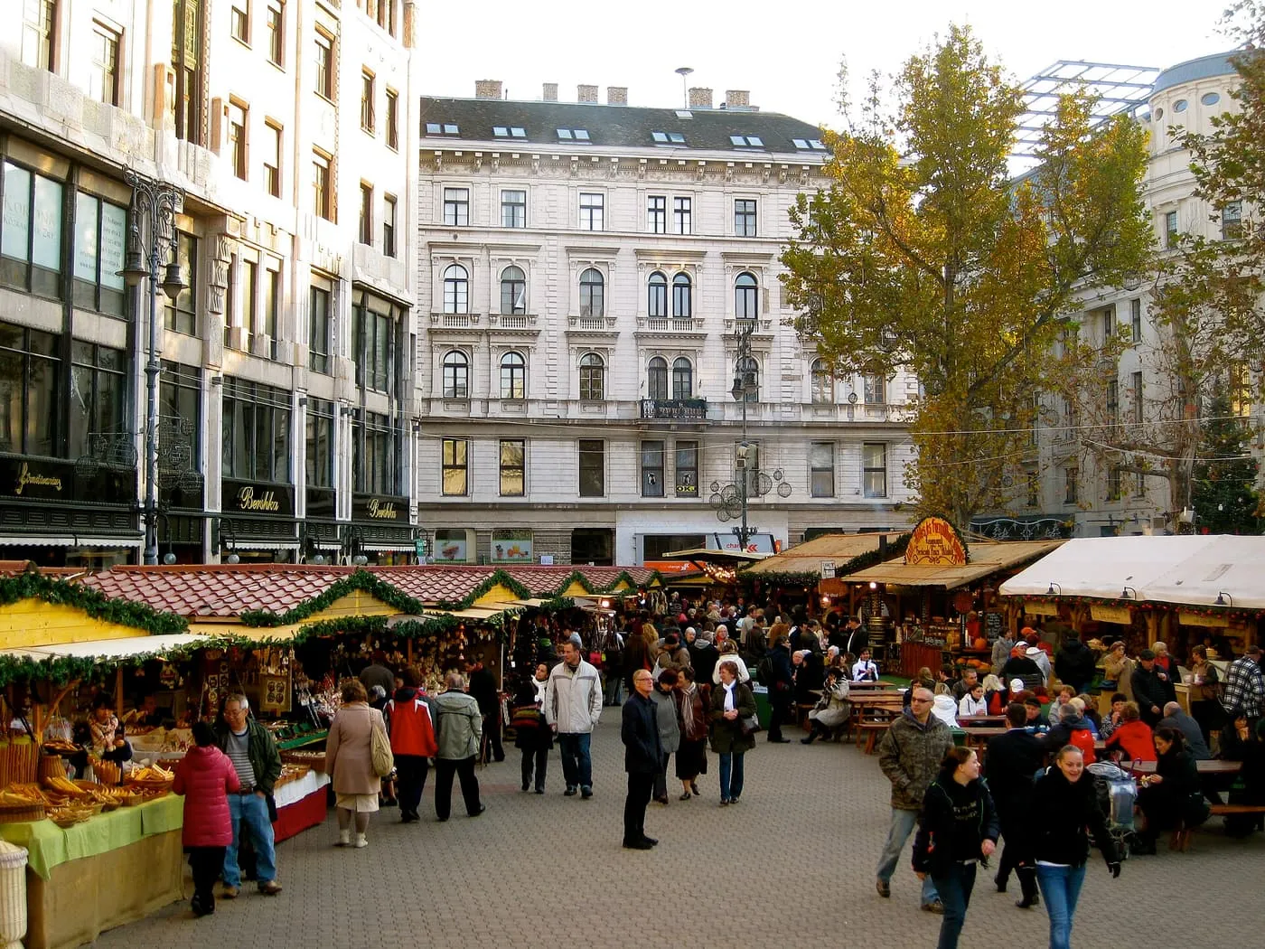 Wooden Christmas market stalls trimmed with greenery on Vörösmarty tér, with visitors walking between them in daylight