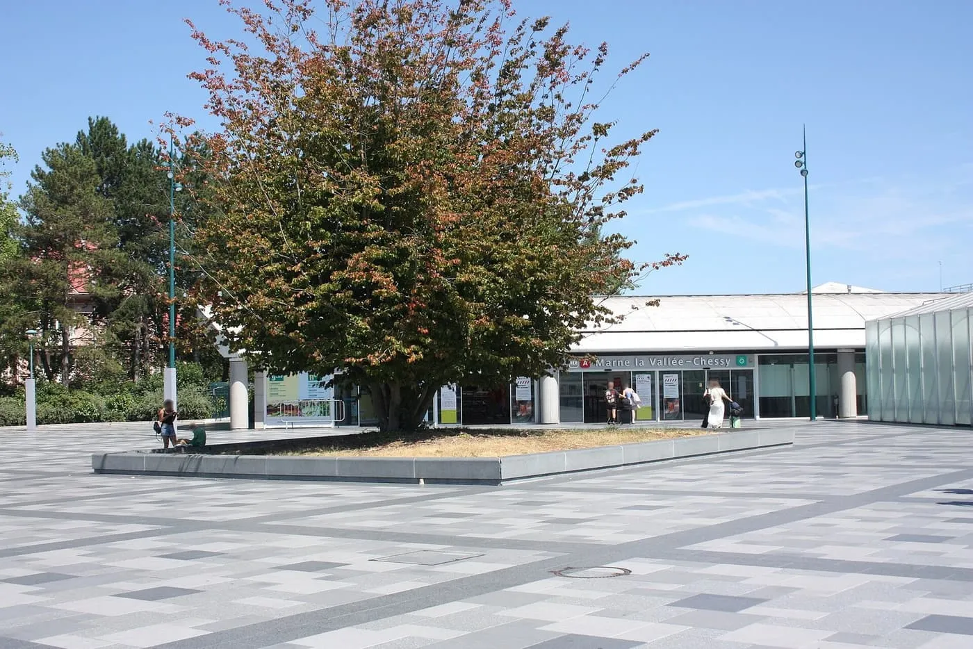 The RER A entrance of Marne-la-Vallée–Chessy station, a low glass-fronted hall behind a paved square with a large tree