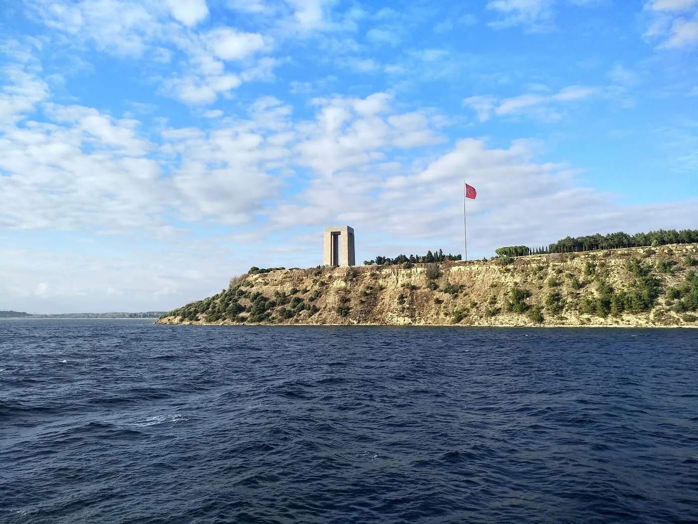 The tall stone Çanakkale Martyrs' Memorial and a flagpole on a low cliff above dark blue water