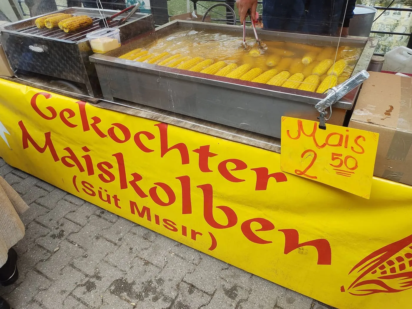A market stall with boiled and grilled corn on the cob behind a yellow banner reading Gekochter Maiskolben