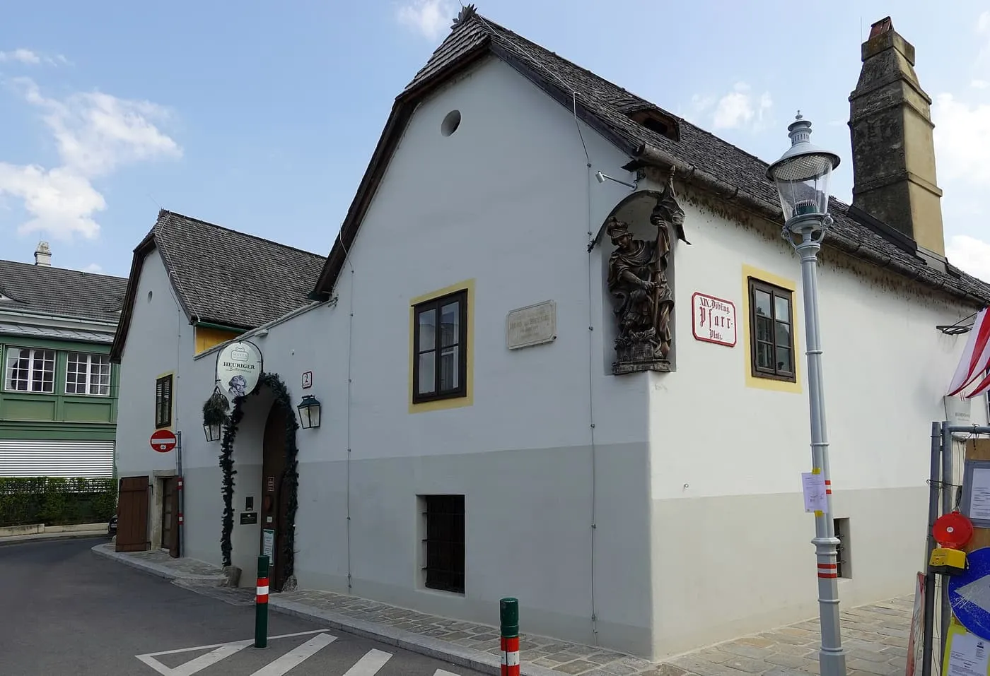 The white and grey gabled house on Pfarrplatz with a Heuriger sign and a wreathed archway, and a carved figure on the corner