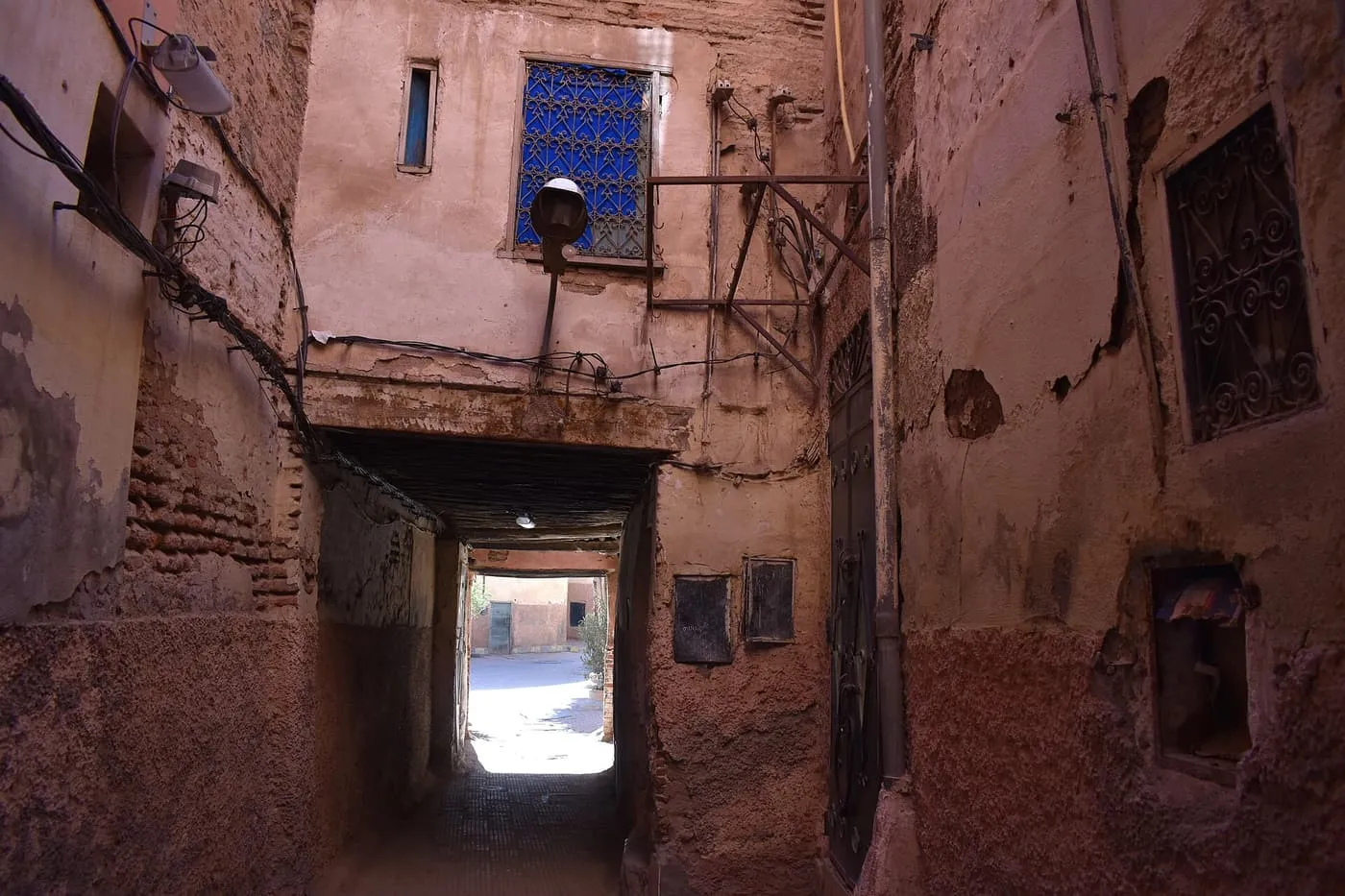 A narrow medina lane passing through an arched tunnel under a house, with a blue lattice window above