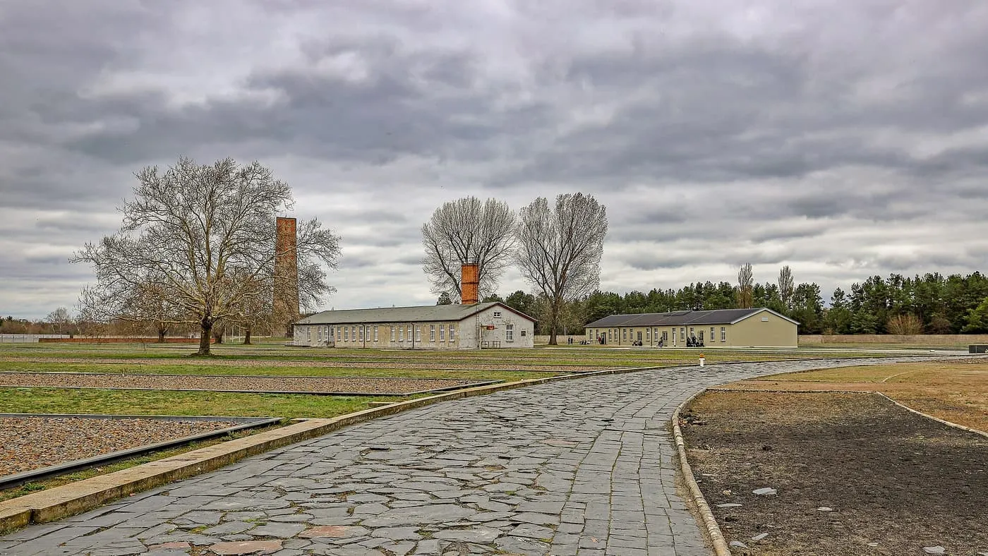 A stone path across the grounds of the Sachsenhausen memorial, with gravel outlines of barracks, bare trees and surviving buildings under a cloudy sky