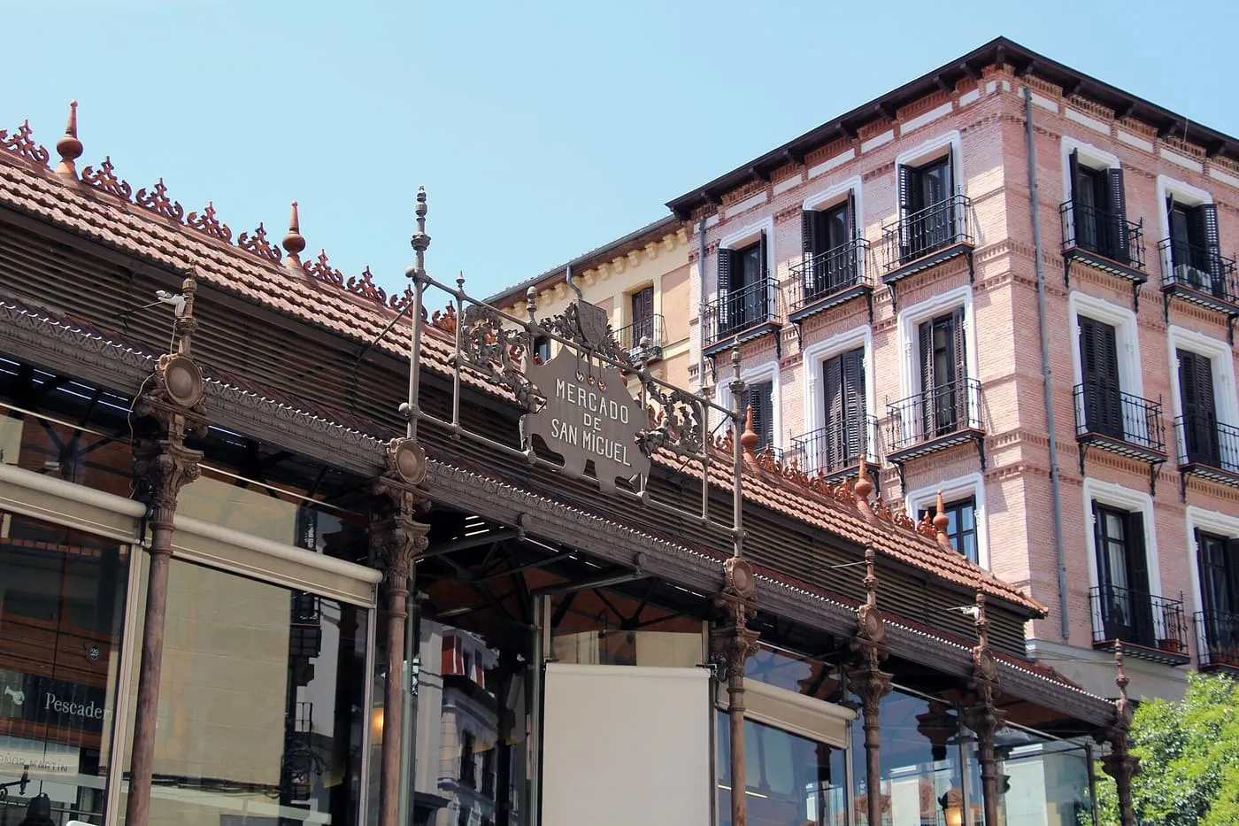 The iron-and-glass front of the Mercado de San Miguel with its tiled roof and the market's name on the ironwork, beside a pink brick building
