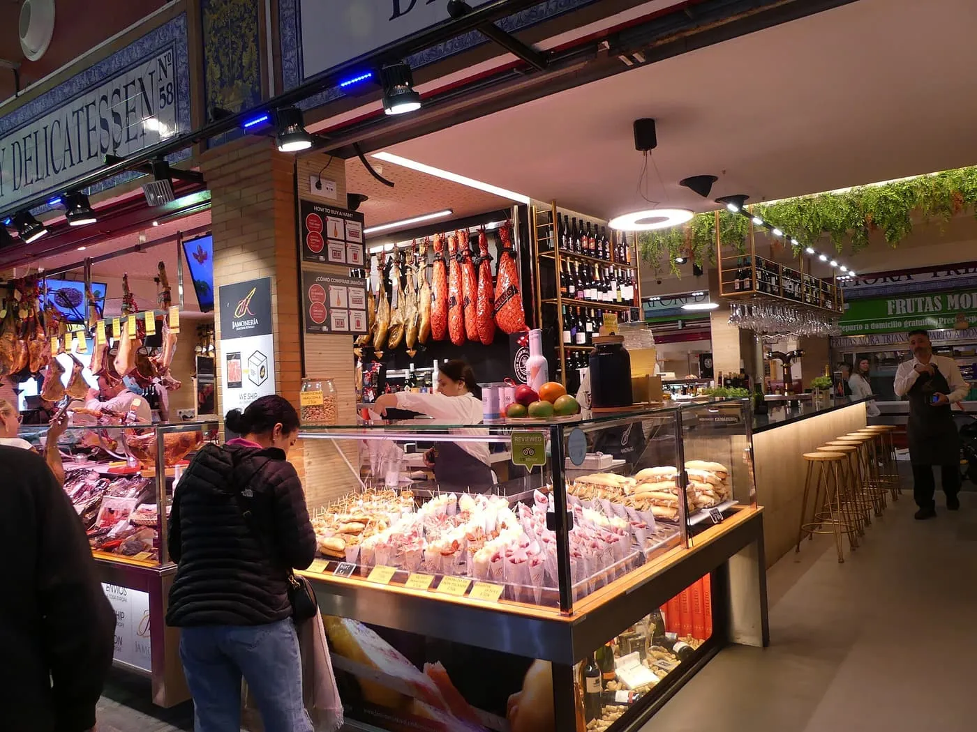 A deli counter inside the Mercado de Triana with legs of ham hanging, a glass case of filled rolls and a customer at the counter
