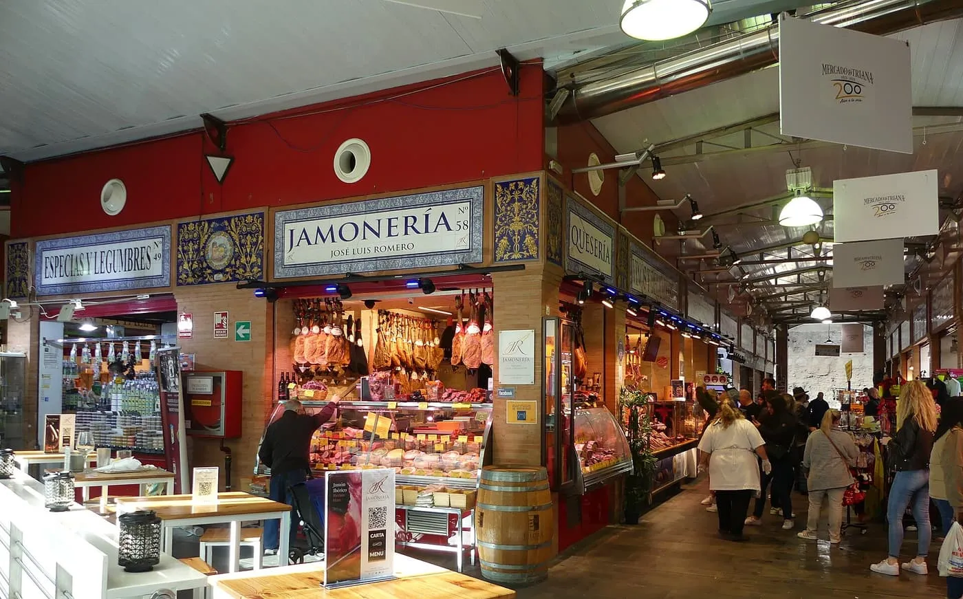 A ham and cheese stall under tiled signs inside the Triana market, with shoppers in the aisle