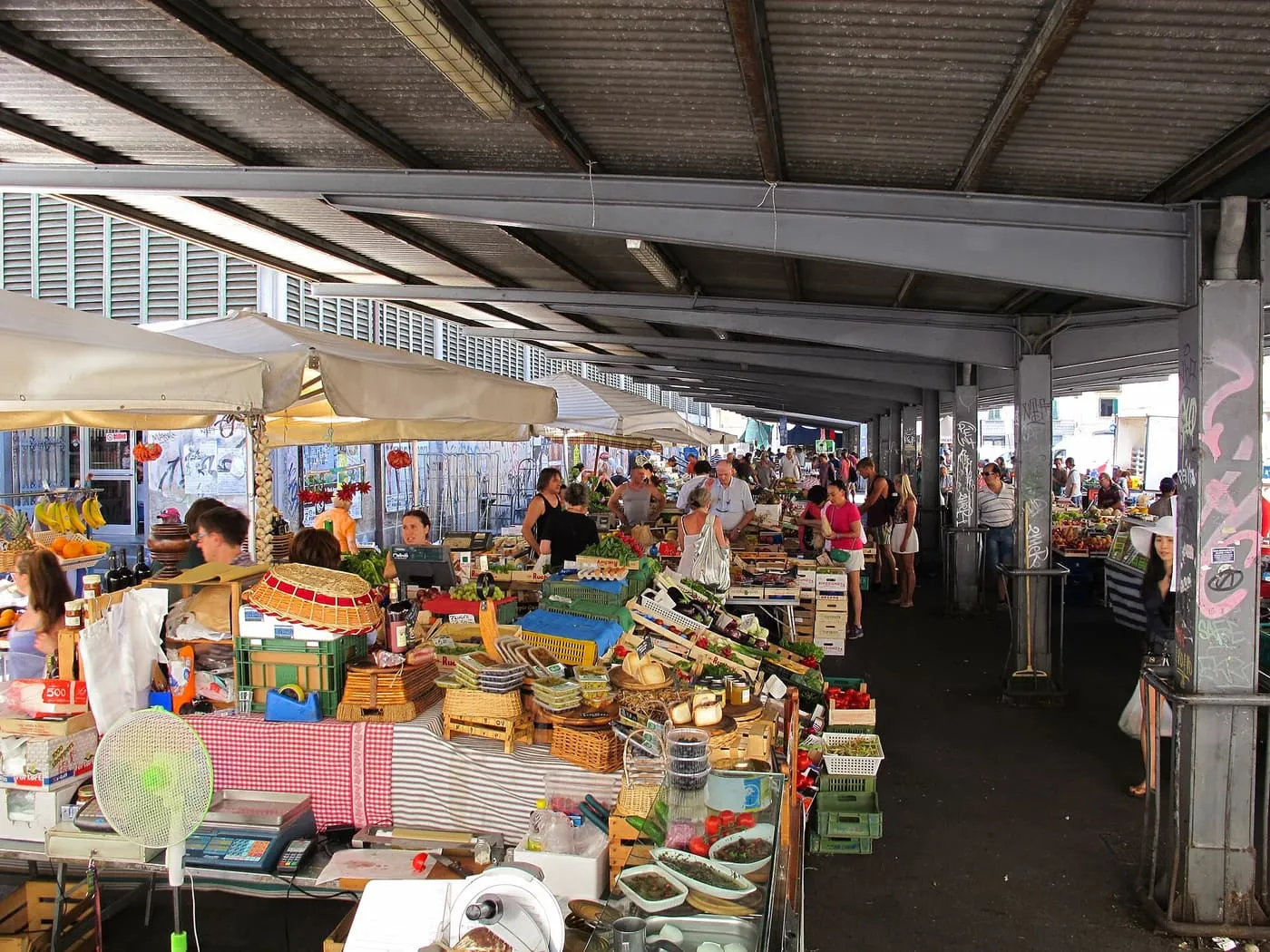Fruit and vegetable stalls under the steel roof of the Mercato di Sant'Ambrogio, with shoppers between the tables