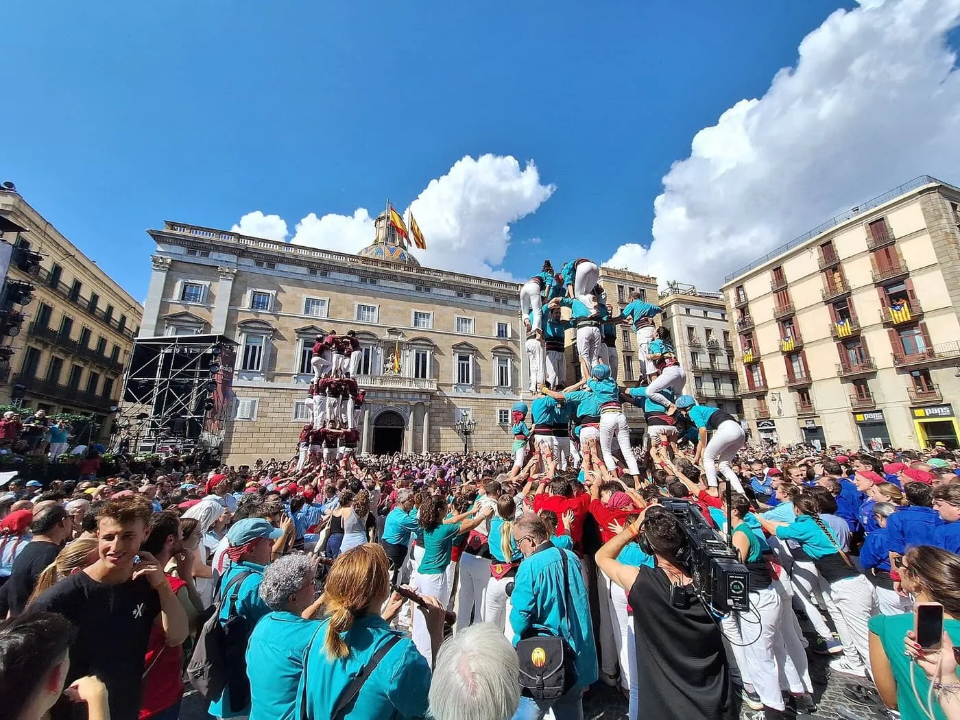 Two human towers rising above a packed Plaça de Sant Jaume, one in teal shirts and one in maroon, in front of the Palau de la Generalitat