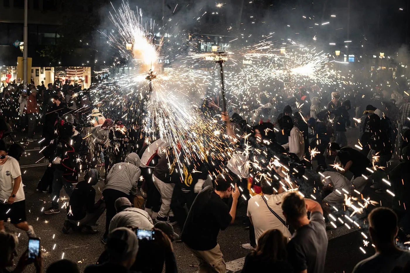 Sparks from a correfoc thrown across a Barcelona street at night, with the crowd crouching and filming through the smoke