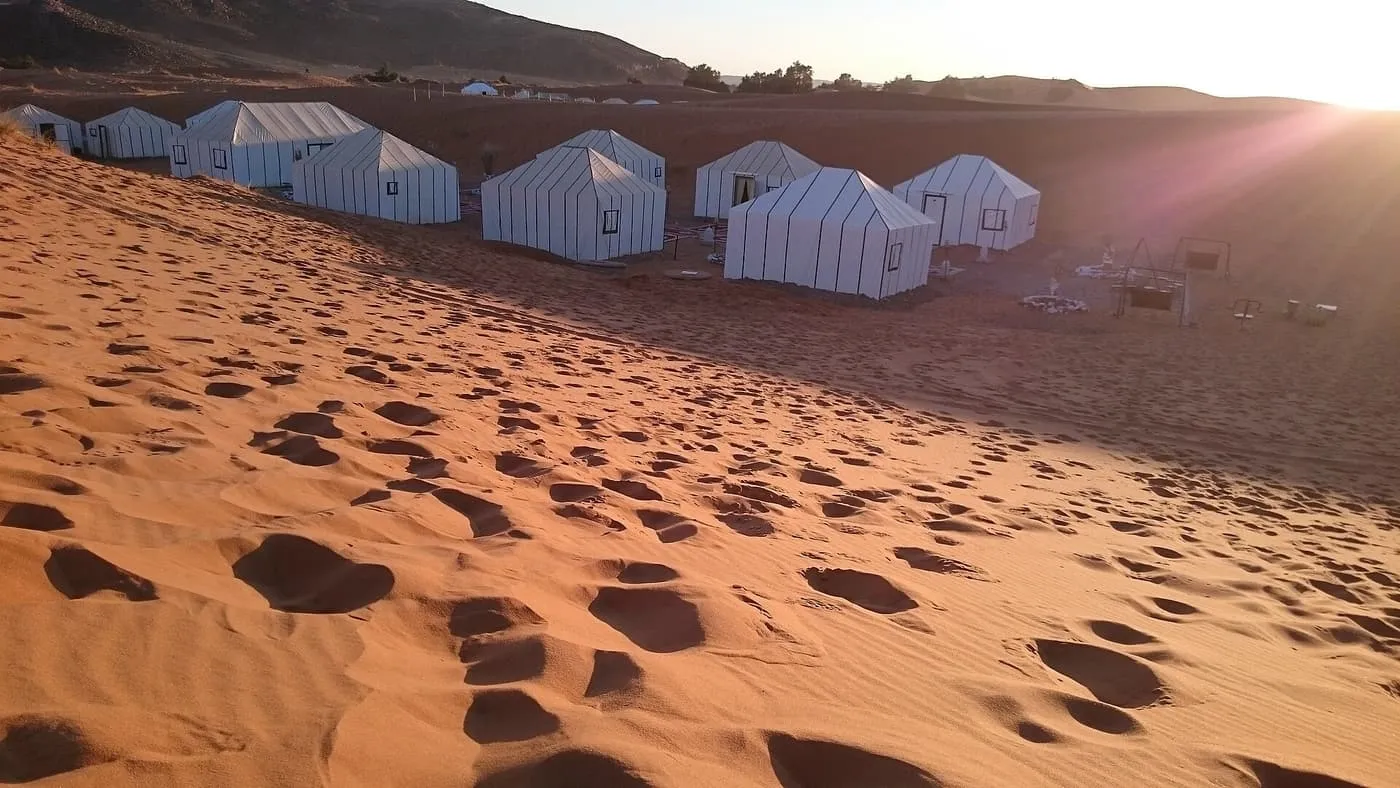 White tents of a desert camp on flat ground below a dune slope marked with footprints