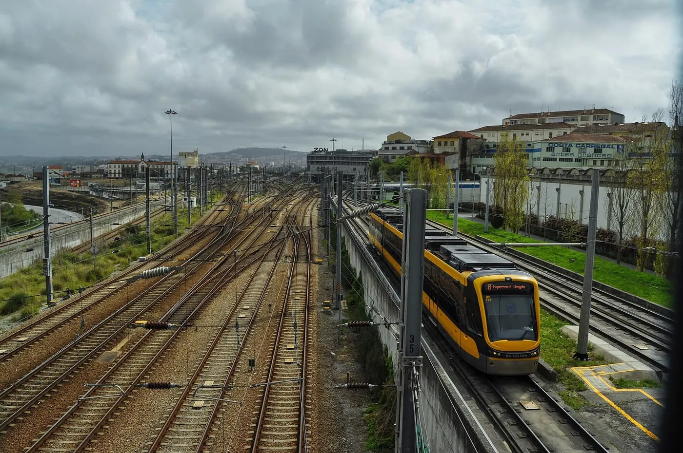 A yellow and grey Metro do Porto tram running beside a wide rail yard at Campanhã under a cloudy sky
