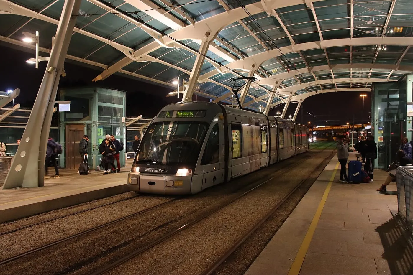 A grey Metro do Porto tram marked E Aeroporto at the airport platform, under a curved glass and white steel roof, at night